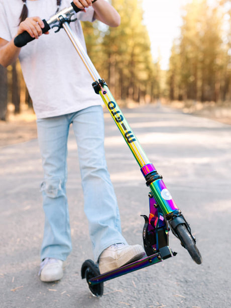 Enfant avec une trottinette Micro Sprite Neochrome aux reflets irisés, pliée sur une route en forêt