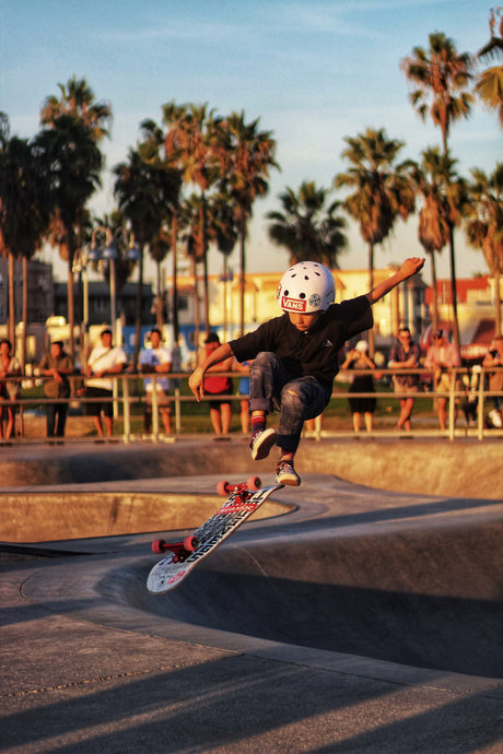 Enfant avec casque faisant un saut en skateboard dans un bowl de skatepark au coucher du soleil